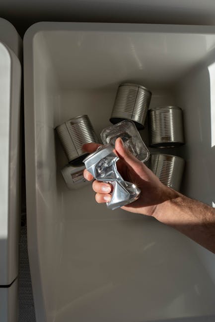 A person's hand is shown placing a crumpled brown paper bag into the open lid of a grey rubbish bin, which is situated on a wooden surface. The person is wearing a white sleeve, and the bag appears to