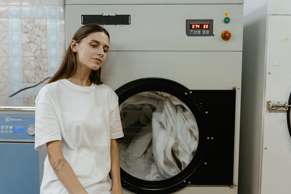 An industrial laundry room featuring a large front-loading washing machine constructed from stainless steel with a circular glass door showing white fabric inside. The machine has a yellow warning sti