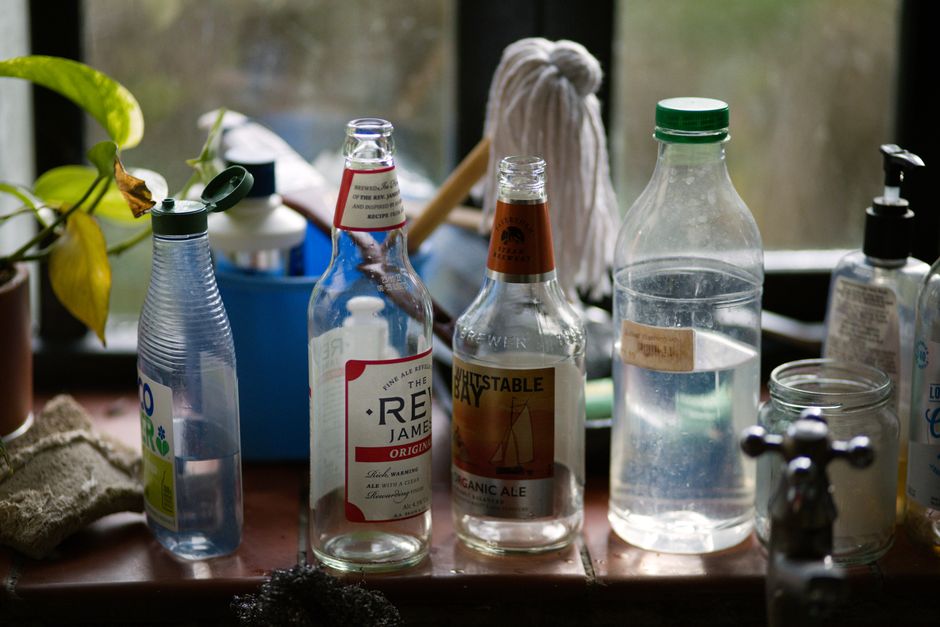 A collection of used glass bottles and containers placed on a wooden surface, situated near a window with natural daylight illuminating the scene. The bottles include a small clear plastic water bottl